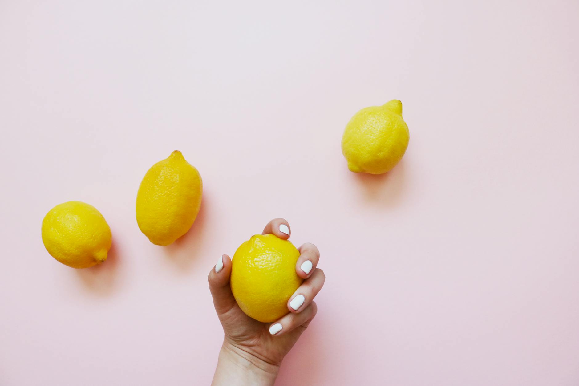 A hand with white nails holding a fresh lemon on a soft pink background surrounded by additional lemons