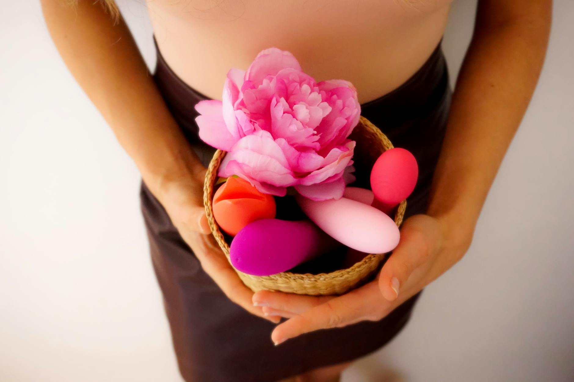 A collection of colorful vibrators and a fresh pink flower in a basket, representing diverse pleasure tools and intimate wellness.